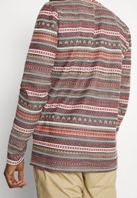Langsleeve shirt met een geometrisch patroon in rood, zwart en beige. Gemaakt van een textiel met textuur, met contrasterende strepen en ontwerpen.