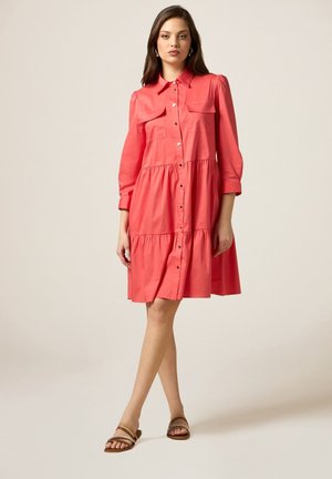 Korallfarbenes Hemdblusenkleid mit Kragen, langen Puffärmel, zwei Brusttaschen, gestuften Rock und glattem Stoff, kombiniert mit braunen flachen Sandalen.
