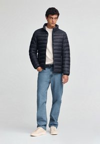 Czarna kurtka puchowa z poziomym pikowaniem, wysokim kołnierzem i czerwonym logo. Nosi się ją na jasnym swetrze, w połączeniu z jasnoniebieskimi dżinsami i jasnymi sneakersami.