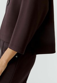 Dunkelbraunes, weiches Stoffoutfit mit einem lockeren Oberteil mit Dreiviertelärmeln und passenden, schmal geschnittenen Hosen, die durch einen dezenten Glanz hervorgehoben werden.