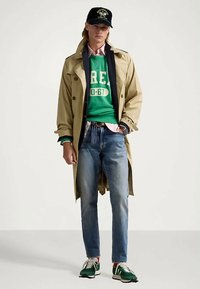 Trench coat bege, sweatshirts verde com impressão branca, jeans azuis e ténis verdes. Os acessórios incluem um boné preto e camisetas em camadas.