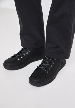 Zapatillas negras de caña alta con parte superior de tela, punta redonda, diseño con cordones y suela de goma. Se usan con pantalones oscuros, con ojales y cordones plateados.