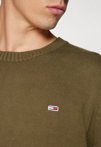 Oliivinvihreä neulepaita pyöreällä kaula-aukolla ja pienellä rintakehään kirjotulla logolla punaisena, valkoisena ja sinisenä. Pinta on teksturoitu ja reunat resoritut.