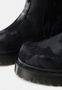 Melni ādas zābaki ar teksturētām kažokādas daļām, biezām ribotām zolēm un sānu rāvējslēdzēju aizdari tuvplānā.