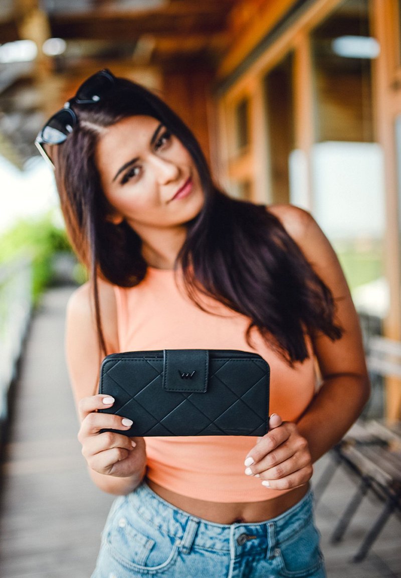 Black leather wallet with quilted pattern and a magnetic clasp, held by a person. Background shows a wooden structure and blurred surroundings.