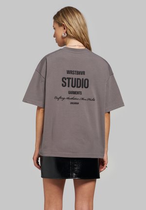 Femme aux longs cheveux portant un t-shirt gris ample avec un texte noir et une mini-jupe noire brillante, vue de dos sur un fond uni.