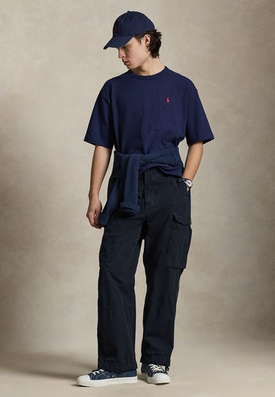 Νεαρός άνδρας που φοράει σκούφο μπλε ναυτικού, μπλε ναυτικό t-shirt με κόκκινο λογότυπο, παντελόνια cargo, πουλόβερ δεμένο γύρω από τη μέση και αθλητικά παπούτσια, κοιτάζοντας προς τα πλάγια.