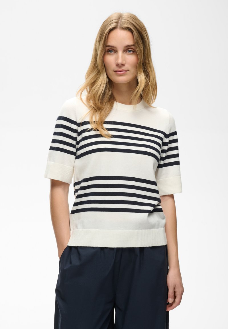 Femme aux longs cheveux blonds portant un pull blanc à manches courtes avec des rayures horizontales noires et un pantalon foncé, debout devant un fond uni.