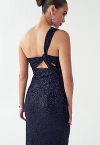 Tumši zila pailleta kleita ar pieguļošu siluetu, aizmugurē ar izgriezumu, plecu siksnās ar pagrieziena detaļu un gludu, mirdzošu tekstūru.