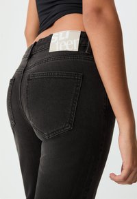 Jeans in denim nero con un design aderente, dotati di tasche posteriori e un'etichetta grigia chiara sulla vita, che mostra un branding minimalista.