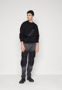 Felpa nera con maniche lunghe, pantaloni cargo grigi con più tasche, marsupio nero e stivali neri. Design semplice su uno sfondo neutro.