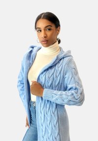 Ljusblå kabelstickad cardigan med huva, med ribbade ärmslut och öppen front, bärs över en beige polotröja och blå jeans.