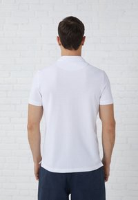 Witte polo met een zachte, gladde textuur, korte mouwen en een klassieke kraag, gecombineerd met blauwe shorts, gezien vanuit de achterkant.