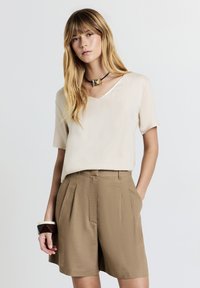 Donna con lunghi capelli biondi che indossa una maglietta beige a maniche corte, pantaloni corti marroni a vita alta, una collana con pendente quadrato e un braccialetto geometrico scuro.