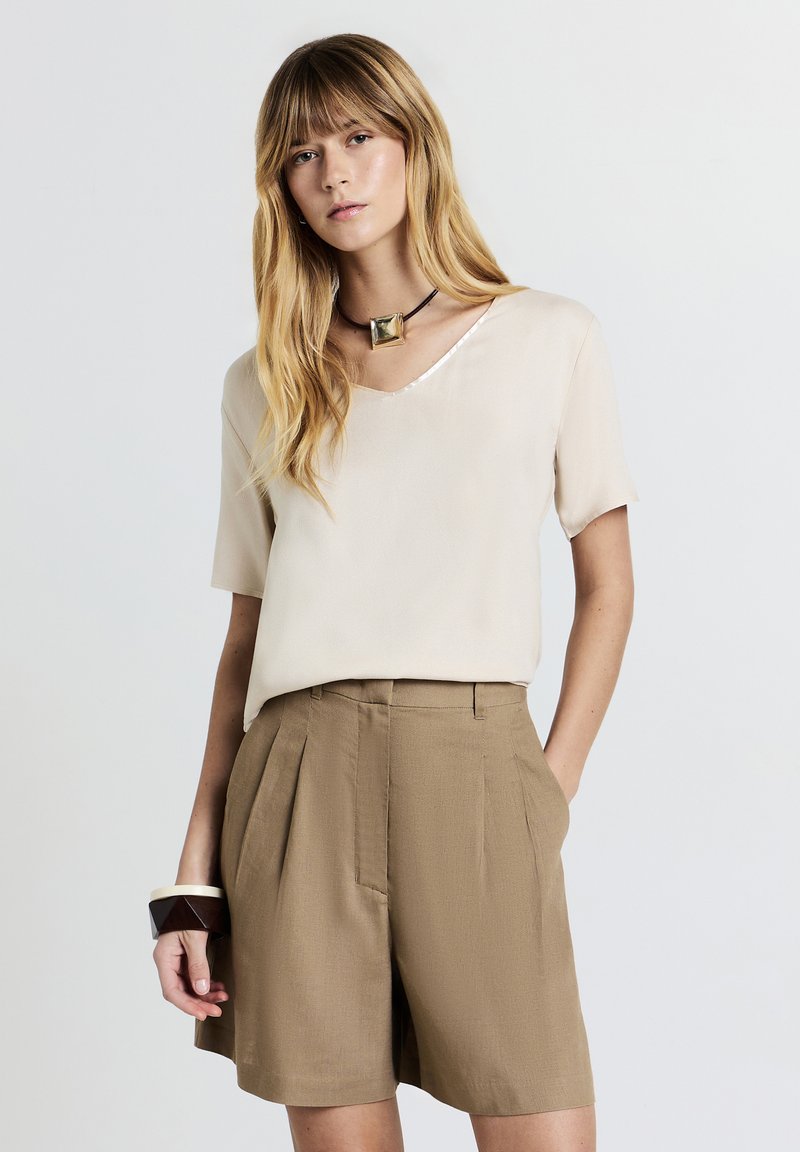 Donna con lunghi capelli biondi che indossa una maglietta beige a maniche corte, pantaloni corti marroni a vita alta, una collana con pendente quadrato e un braccialetto geometrico scuro.