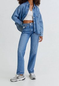 Giacca di jeans azzurra chiara e jeans a vita alta, abbinati a un top corto bianco e sneakers grigie, che mostrano una vestibilità rilassata e uno stile casual.
