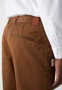 Brown cotton pants feature a label at the waistband, a side pocket with a decorative stitch, and a smooth texture.