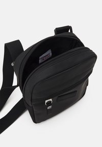 Borsa a tracolla nera realizzata in nylon liscio, con chiusura a zip, tracolla regolabile e un discreto patch logo sul davanti. L'interno è non foderato.