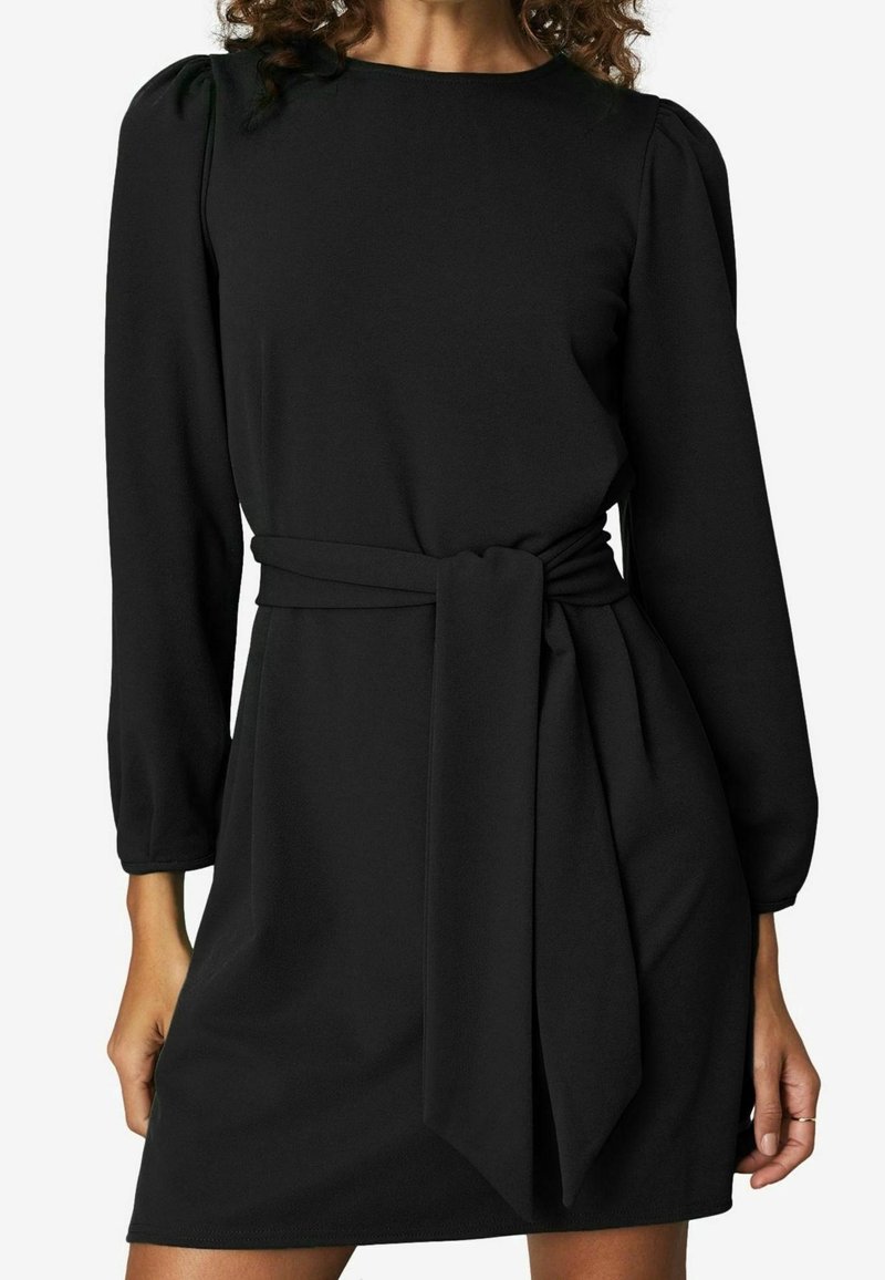 Femme portant une robe noire à manches longues avec des épaules bouffantes et une large ceinture en tissu nouée à la taille.