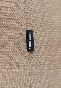 Beidzis adīts audums ar mazu melnu taisnstūra birku, kas ir piešūta, uz kuras redzams zīmola nosaukums "JACK & JONES" baltām burtām.