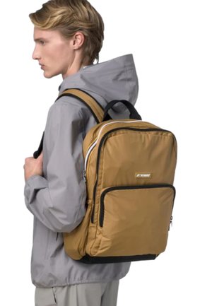 Giovane persona che indossa una giacca con cappuccio grigio chiaro e porta uno zaino grande color beige con bordi neri e dettagli bianchi delle cerniere.