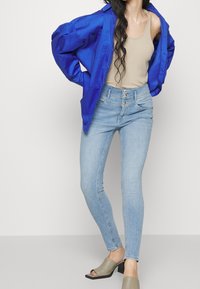 Ljusblå skinny jeans med hög midja och femknappsstängning, kombinerat med en beige linne och en överdimensionerad blå jacka, samt gröna klacksandaler.