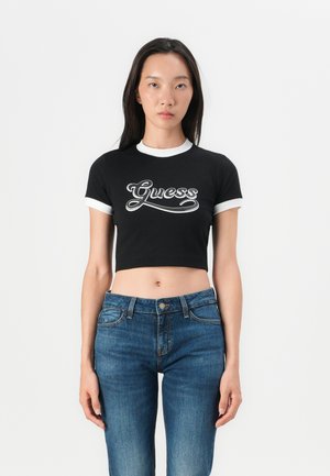 Black cropped t-shirt with white ribbed collar and sleeves. Features white "Guess" logo in cursive across the front. Worn with blue denim jeans.
