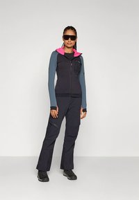 Schwarze und graue Sportjacke mit pinkfarbener Kapuze, kombiniert mit schwarzer Hose. Merkmale sind ein Reißverschluss vorne, Daumenlöcher und Taschen. Geeignet für den Außenbereich.