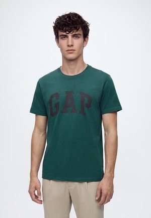 Ung mand med krøllet mørkt hår iført en grøn GAP T-shirt og beige bukser, der står mod en ensfarvet lys baggrund.