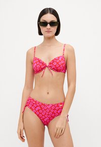Rožinė bikini maudymosi kostiumėlis su gėlių raštais ir tekstiniais akcentais, su reguliuojamais dirželiais ir surišimu centre viršaus dalyje. Minkšta tekstūra.