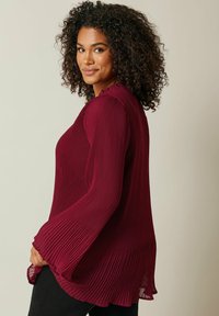 Blouse plissée bordeaux avec de longues manches évasées. Présente un tissage texturé et un col festonné. Tissu lisse avec une coupe décontractée.