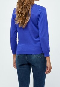 Femme aux longs cheveux ondulés portant un pull ajusté bleu cobalt et un jean bleu foncé, debout devant un fond clair uni.