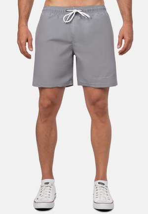 Uomo che indossa pantaloncini da bagno grigi con coulisse e sneakers bianche basse, in piedi davanti a uno sfondo semplice.