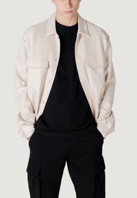 Beige button-up jack met twee borstzakken, gedragen over een zwart shirt. De jack heeft een relaxte pasvorm en een soepele textuur. Zwarte broek met zakken.