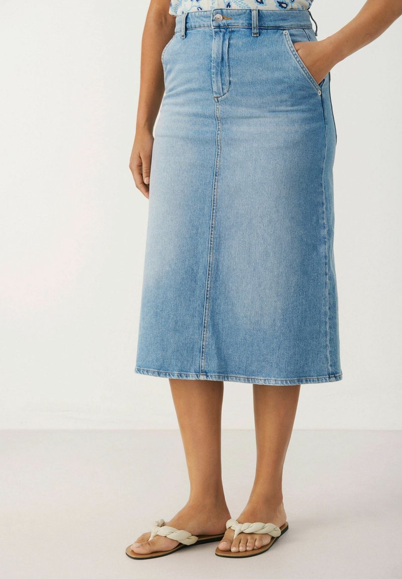 Femme portant une jupe midi en denim bleu clair avec des poches avant, associée à des sandales à brides tressées blanches, debout sur un fond uni.