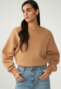 Vaaleanruskea sweatshirt rentoa istuvuutta, ribbadattu pyöreä kaula-aukko ja pudotetut olkapäät, yhdistettynä korkeavyötäröisiin sinisiin farkkuihin.