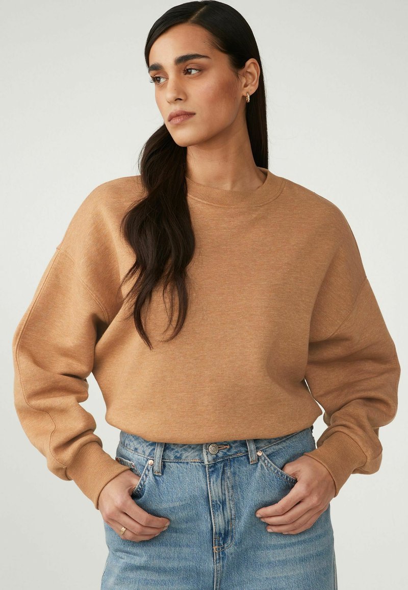 Vaaleanruskea sweatshirt rentoa istuvuutta, ribbadattu pyöreä kaula-aukko ja pudotetut olkapäät, yhdistettynä korkeavyötäröisiin sinisiin farkkuihin.