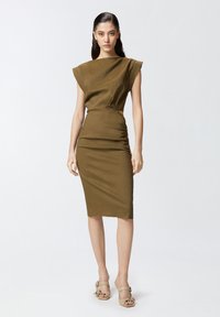 Maslinasto zelena haljina s naglašenim ramena, bočnim izrezima i mekim teksturiranim materijalom. Uparena s sandalama svijetlih boja s kopčama.