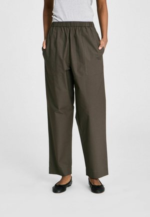 Naine, kellel on seljas laiad, kõrge vöökohaga tumeda oliivivärvi püksid ees taskutega, mustade baleriinikingade ja helehalli ülaosaga.