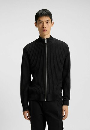 Man wearing a black ribbed knit zip-up sweater with a high collar and black pants, standing against a plain light background.