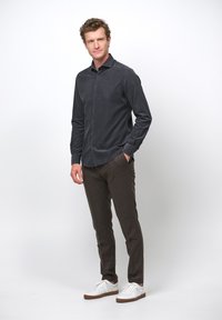 Camicia button-up grigio scuro con una trama liscia, abbinata a pantaloni marroni con texture e sneakers bianche. Il modello si trova davanti a uno sfondo semplice.