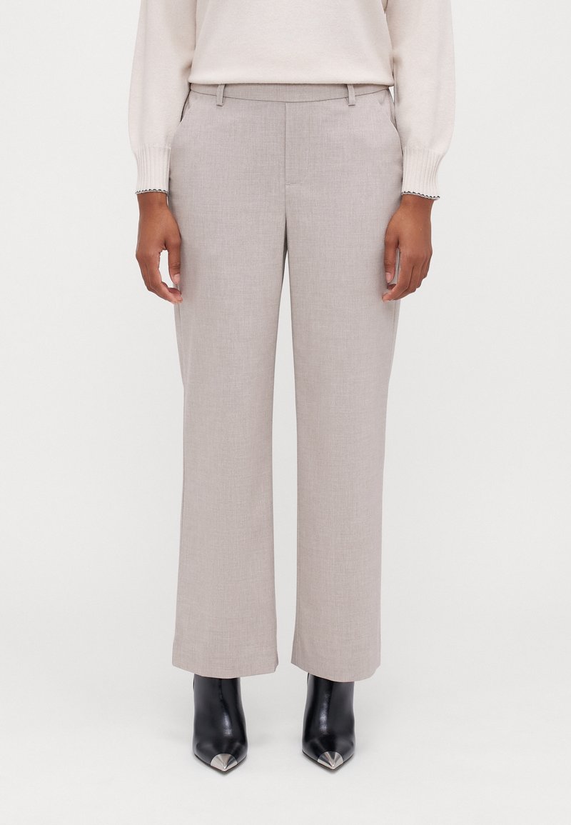 Pantalons larges gris clair en tissu texturé, dotés d'une coupe ajustée, de passants de ceinture et de poches latérales, portés avec des escarpins noirs à bout pointu.