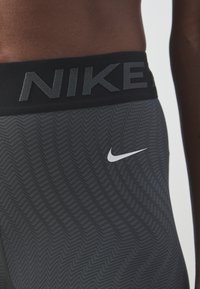Legginsy Nike z czarnym pasem w talii, na którym znajduje się wypukłe logo. Główny materiał jest szary w zygzakowaty wzór oraz posiada białe logo Swoosh.