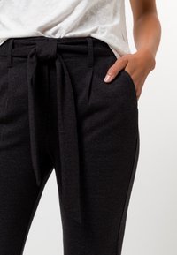 Pantalon noir taille haute en tissu texturé, avec une ceinture à nouer à l'avant et des poches latérales, présenté de la taille aux pieds.