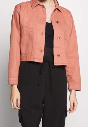 Kvinde iført en kort, falmet lyserød jakke med trykknapper over en sort top og sorte bukser med snøre.