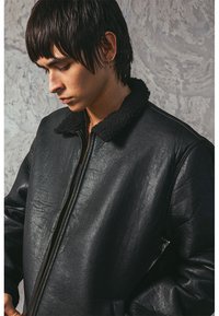 Black leather bomber jacket with a fluffy collar, featuring a front zipper, side pockets, and a smooth texture. Subtle stitching detail.