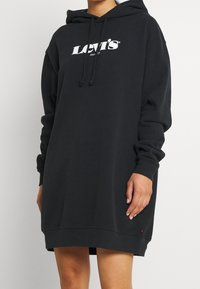 Robe à capuche noire en tissu doux, avec un logo imprimé sur le devant, une capuche à cordon, des manches longues et une coupe décontractée.