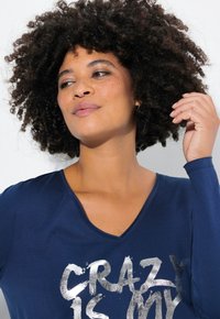 Marineblauwe T-shirt met lange mouwen en V-hals, voorzien van zilverkleurige tekst "CRAZY IS MY" op de voorkant. Zachte stof met een ontspannen pasvorm.