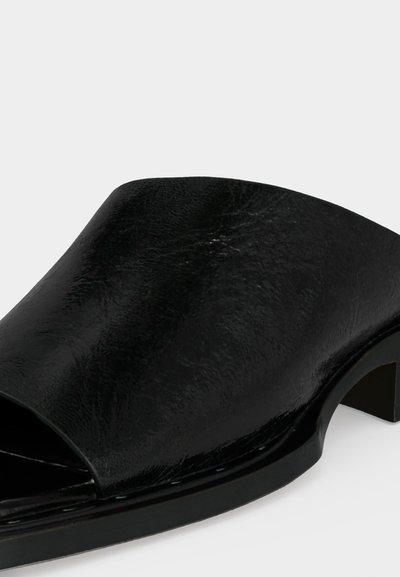 Μαύρο mule παπούτσι με ανοιχτό δάκτυλο, χοντρό χαμηλό τακούνι και λείο δερμάτινο άνω μέρος σε λευκό φόντο.