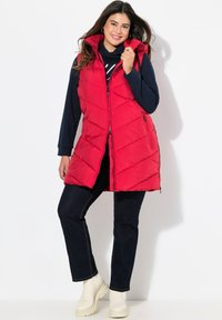 Rote Pufferweste mit hohem Kragen, Reißverschluss und seitlichen Taschen. Kombiniert mit dunklen Jeans und cremefarbenen Stiefeln. Texturiertes gestepptes Muster.